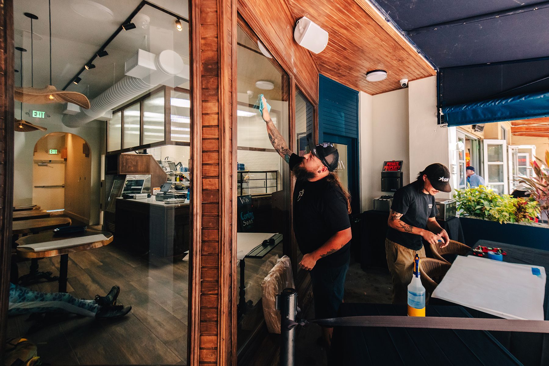 Two men working inside a cafe with glass windows. One cleans the window, another works on a surface, plants and tables are visible.