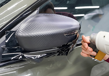 Applying carbon fiber wrap to a car's side mirror with a heat gun, in a workshop setting.