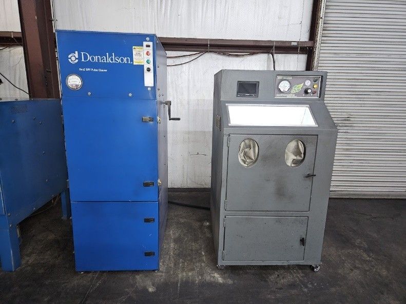 A blue Donaldson industrial dust collector stands beside a gray sandblasting cabinet in a shop setting.