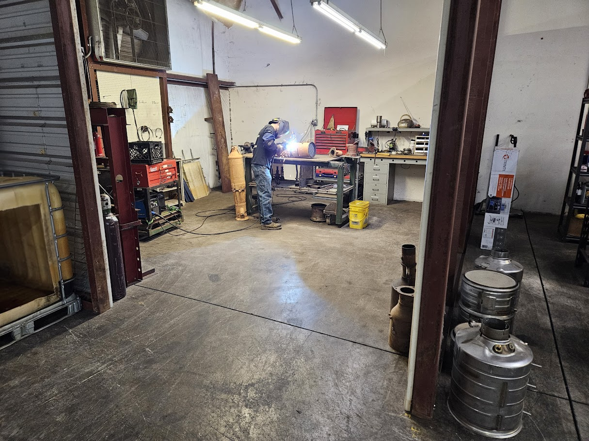 A person in protective gear welds metal at a workbench inside an industrial workshop filled with tools and equipment.