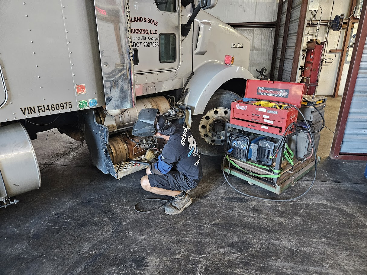 A mechanic wearing a welding helmet works on the side of a parked semi-truck near a red portable tool cart.