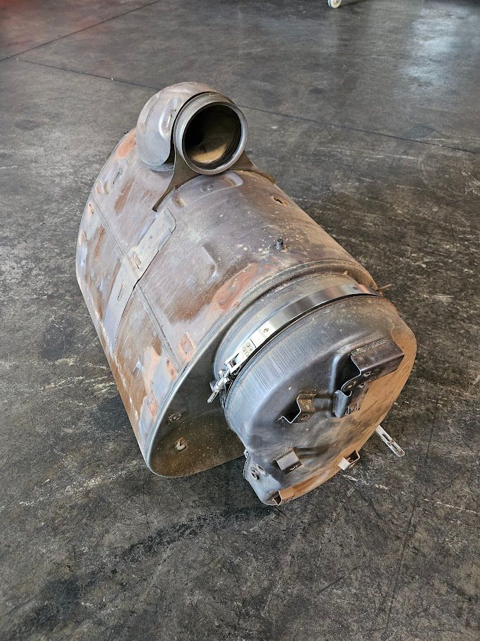 A cylindrical metal heater or wood stove component sits on a concrete floor, featuring a side vent and a circular door.