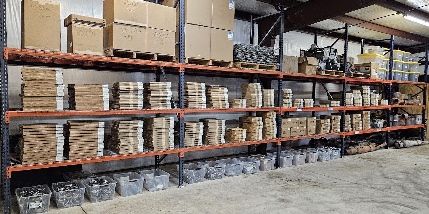 Warehouse shelving units filled with cardboard boxes, stacks of materials, and clear storage bins on a concrete floor.