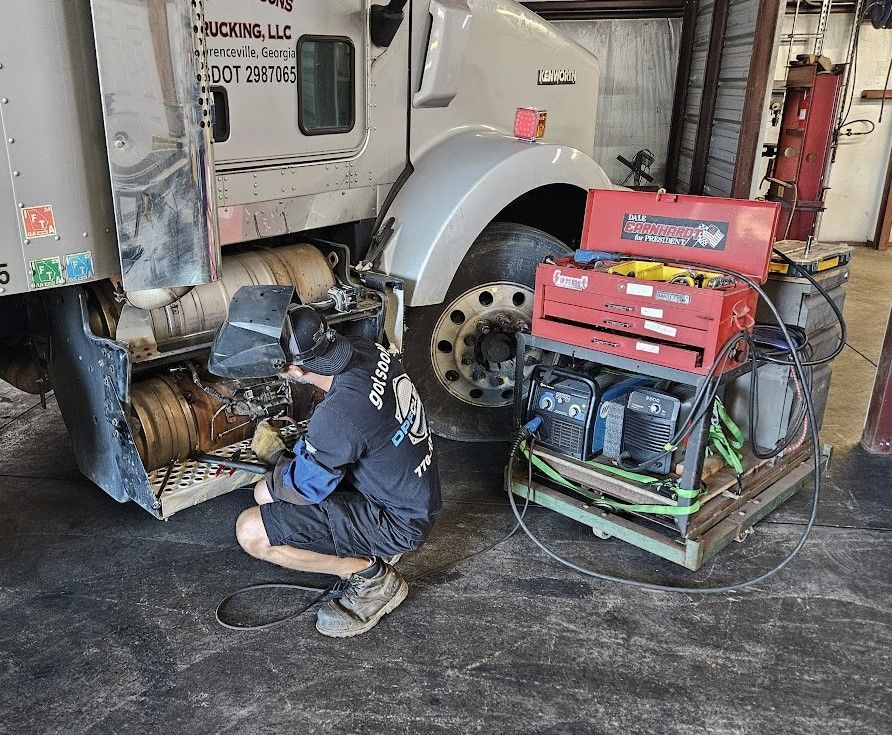 A mechanic wearing a welding helmet crouches to work on the undercarriage of a semi-truck next to a red tool chest.