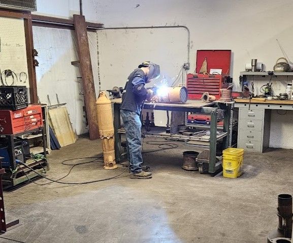 A person wearing a welding helmet welds a metal component at a workbench inside a cluttered workshop.