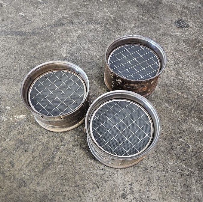 Three circular metal catalytic converter filters with a dark, grid-patterned mesh interior resting on a concrete floor.
