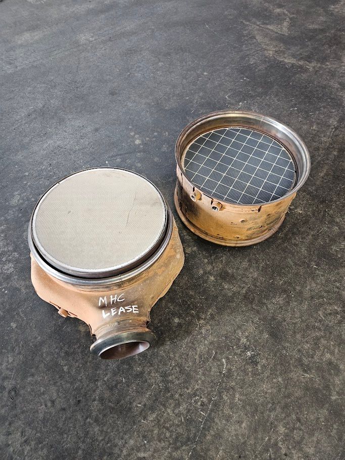 Two round industrial filters on a dark concrete floor; one is covered in light dust, the other has a black wire mesh top.