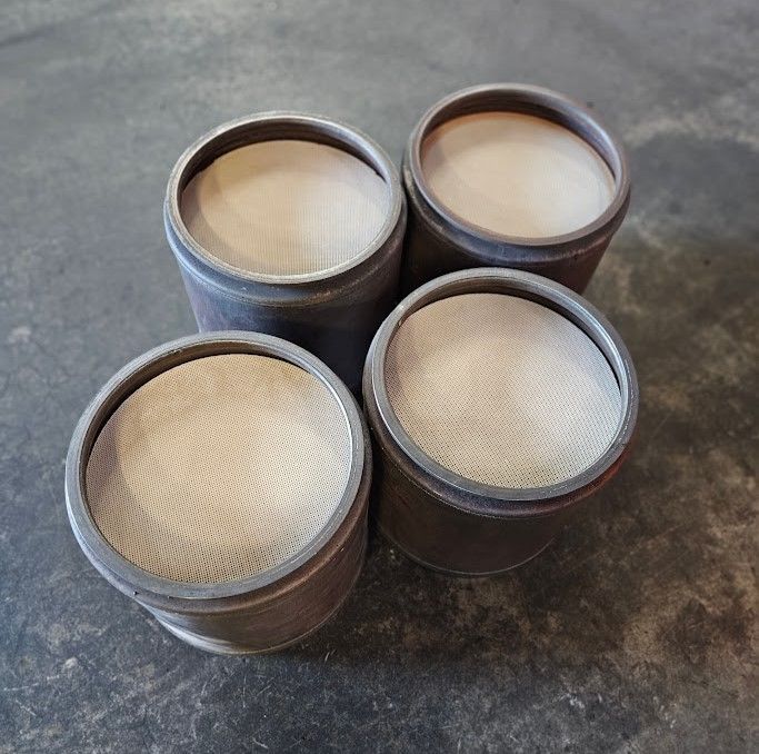 Four cylindrical metal catalytic converter cores with light-colored, honeycomb-patterned ceramic filters on a dark floor.