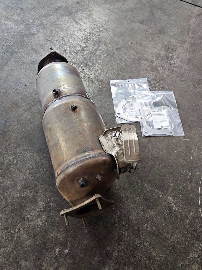 A metal catalytic converter and two small bags containing replacement gaskets sitting on a gray concrete floor.