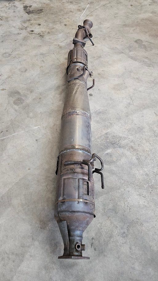 A rusty, cylindrical automotive diesel particulate filter (DPF) with metal pipe connections, lying on a concrete floor.