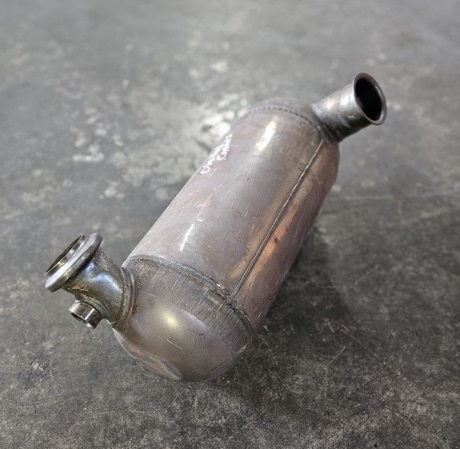 A metallic automotive catalytic converter with two offset pipe connections sits on a dark, textured surface.