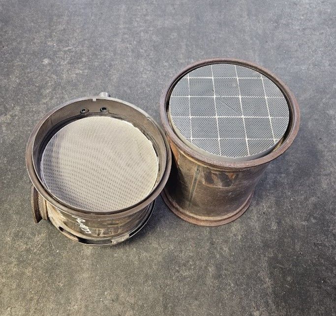 Two cylindrical vehicle catalytic converters with exposed metal honeycomb filter cores lying on a grey surface.