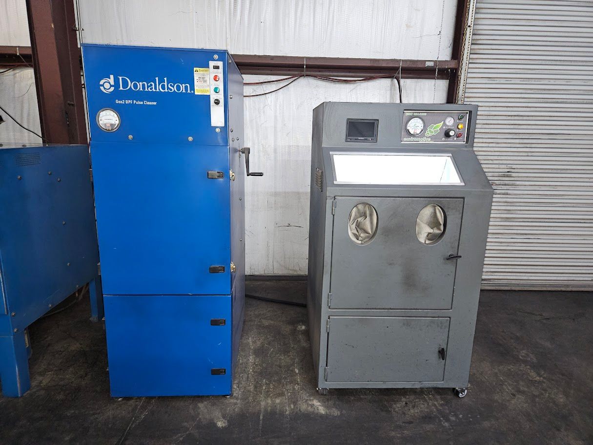 A blue Donaldson dust collector stands next to a gray sandblasting cabinet in an industrial workspace.