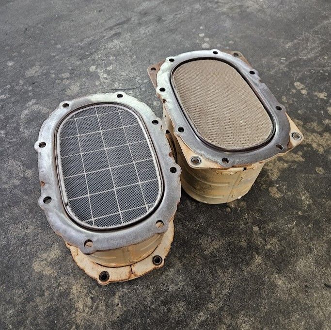 Two oval-shaped car catalytic converters, showing their internal honeycombed ceramic filters, sit on a gray concrete floor.
