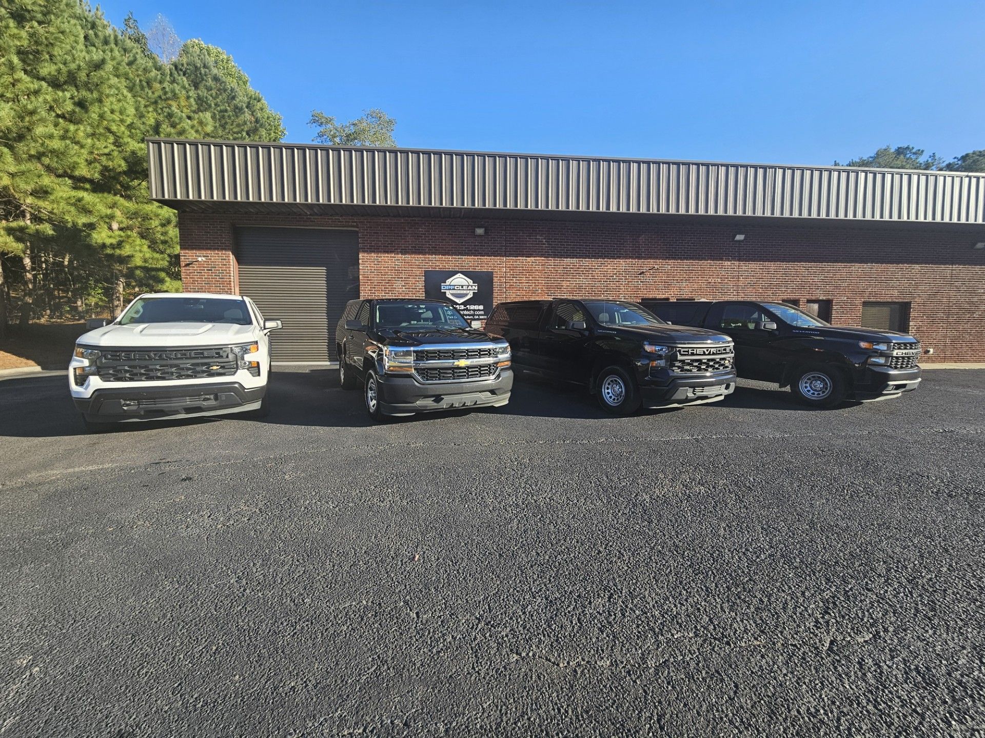 Four trucks parked in a gravel lot in front of a brown, rectangular building on a sunny day.