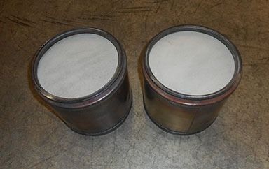 Two cylindrical catalytic converter cores with metallic outer shells and porous white ceramic filter surfaces.