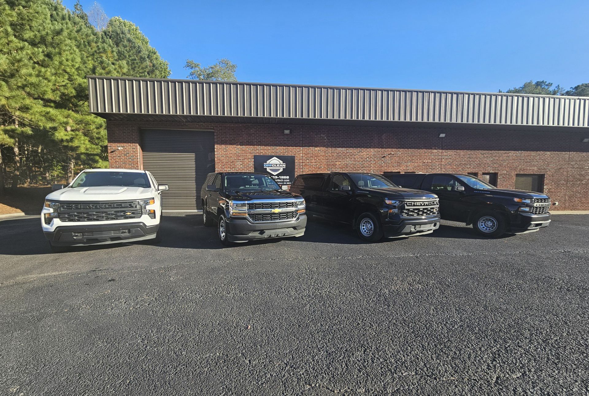 Four pickup trucks, one white and three black, are parked in a row on a gravel lot in front of a brick warehouse.