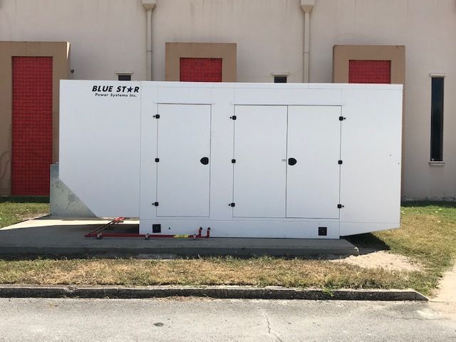 White industrial generator on a concrete pad beside a beige building, with red shutters and grass nearby