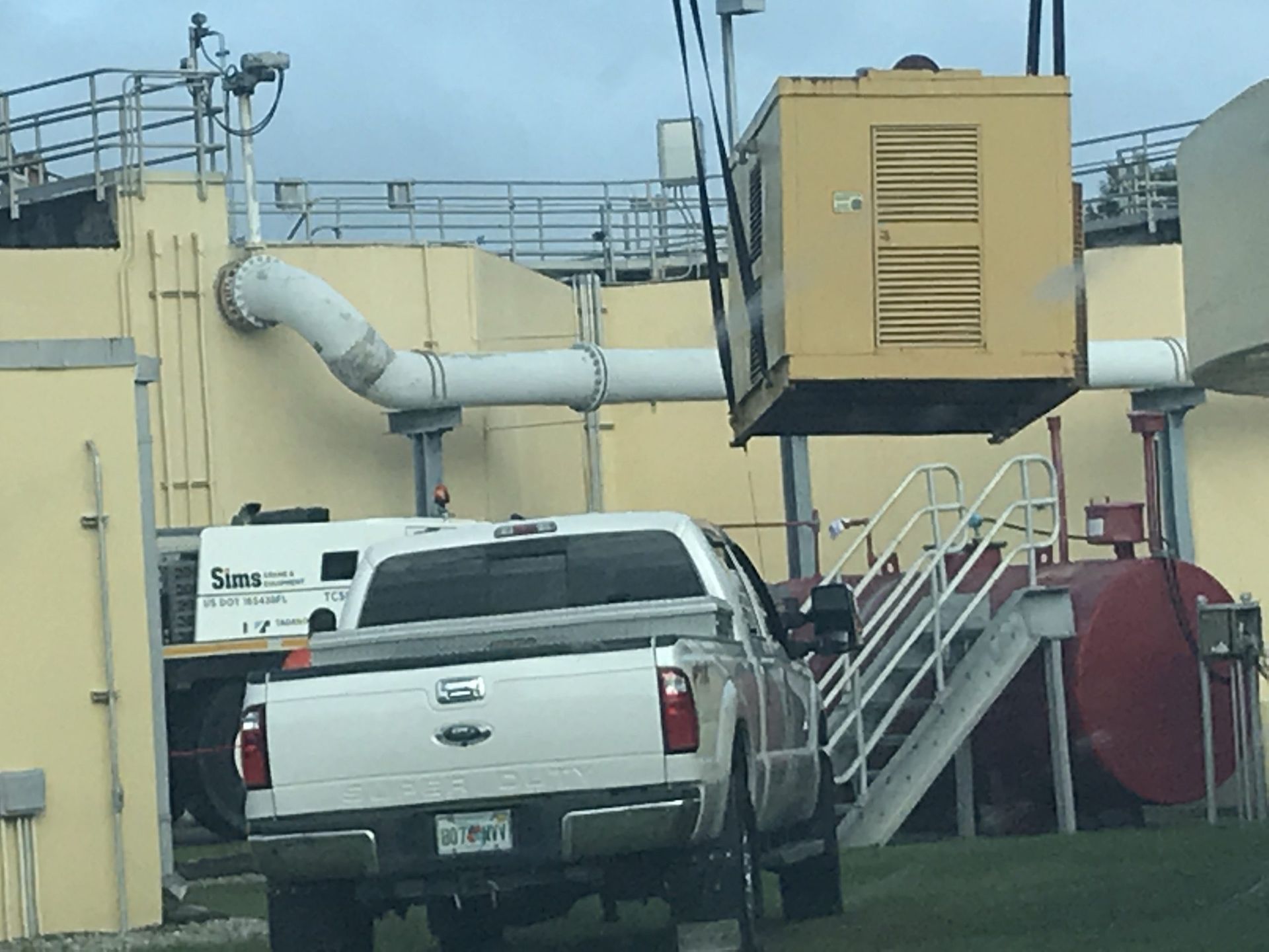 White pickup truck parked beside industrial tanks, pipes, and a yellow utility structure.
