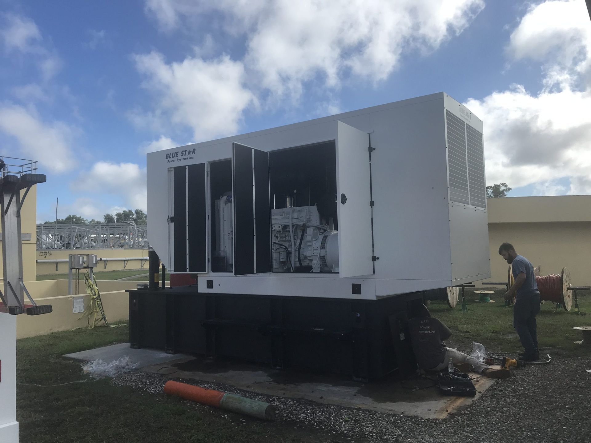 White industrial generator on a trailer in a grassy field, with a person standing nearby under a partly cloudy sky