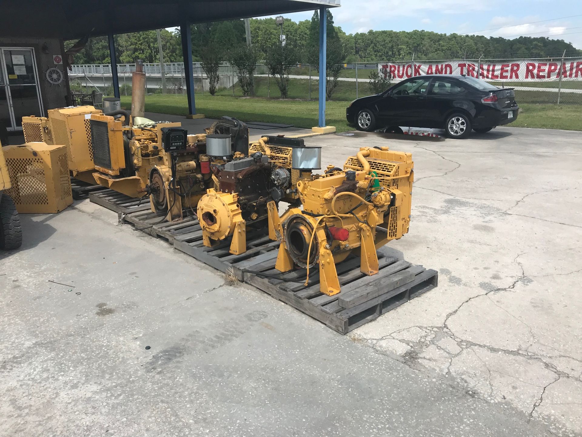 Yellow industrial machinery on pallets beside a black car in an outdoor lot.