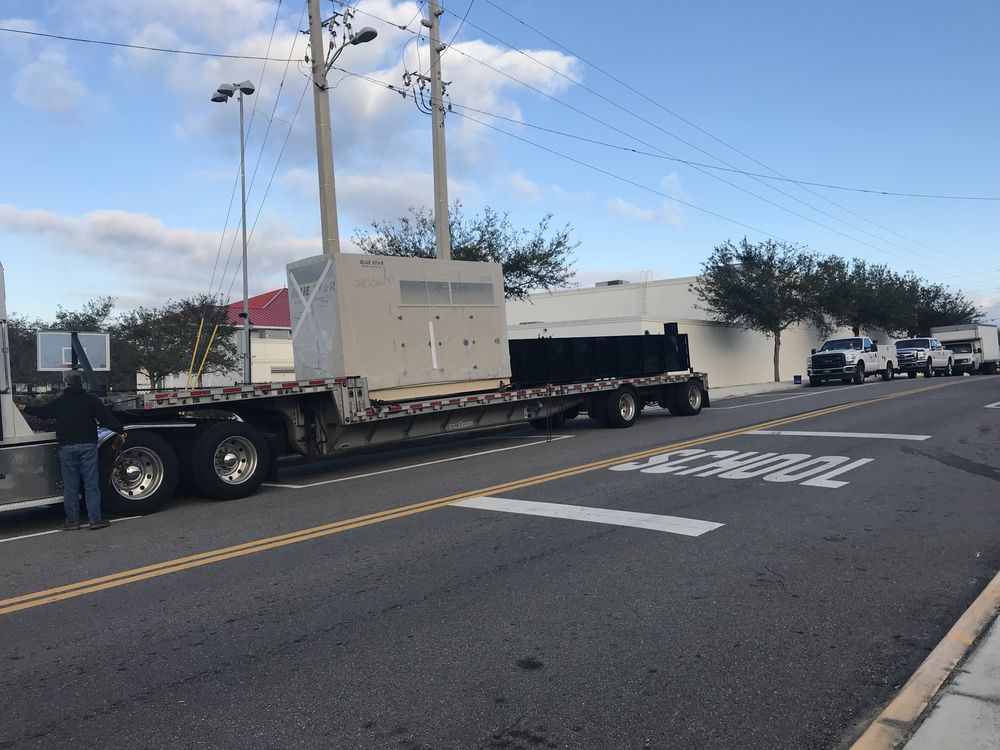 Flatbed truck hauling a large beige machine on a city street with traffic cones and a worker nearby