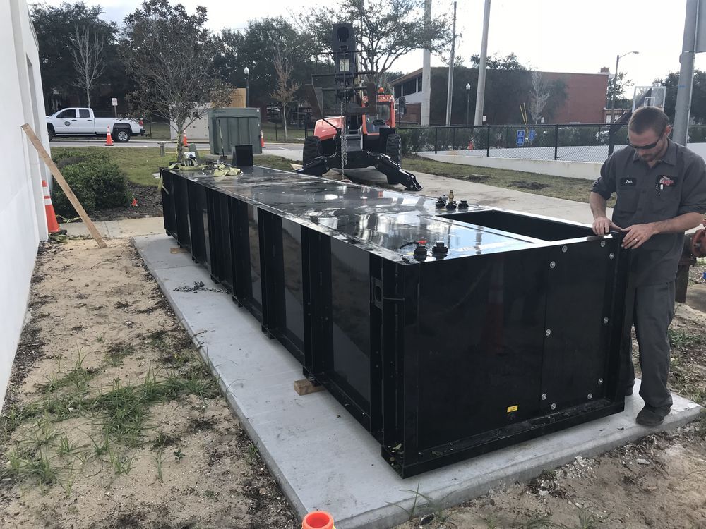 Workers setting a large black metal container on a concrete pad outdoors.