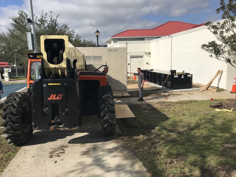 Large JLG telehandler on a construction site beside a white building and paved path under cloudy skies