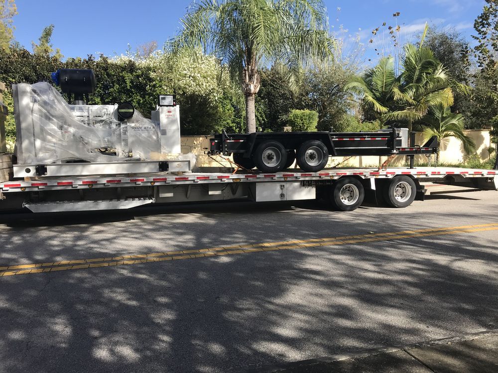 Flatbed trailer hauling a white equipment machine parked on a sunny street with palm trees.