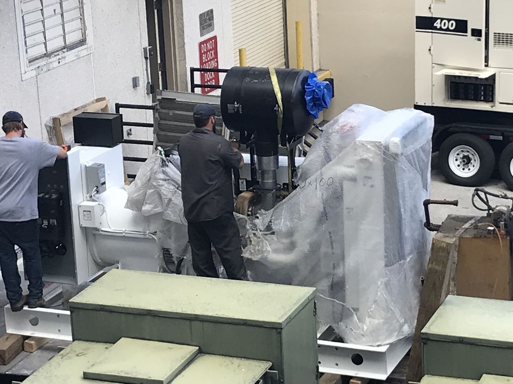 Workers loading a large machine wrapped in plastic onto a truck in a warehouse-like area