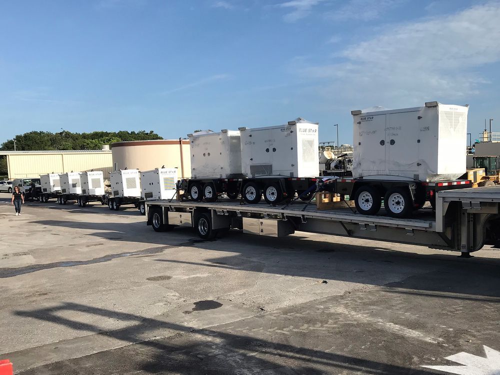 Flatbed trailer carrying several white industrial generators parked in a sunny lot.