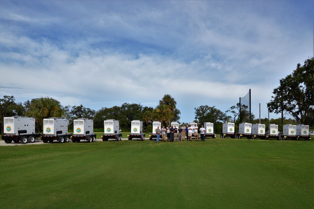 Row of white food trucks on a grassy field under a blue sky, with people gathered near the center