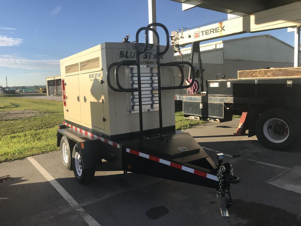 Beige utility trailer-mounted machine parked outside a building with a black hitch and ladder rack.