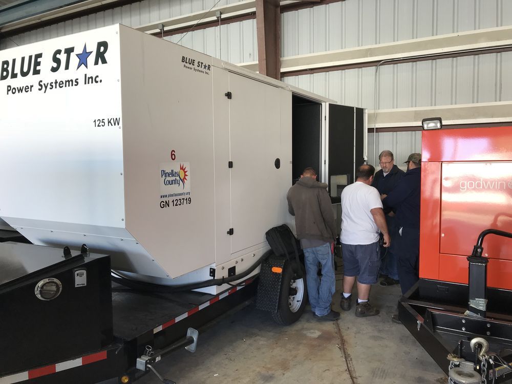 People inspecting a large white Blue Star generator beside an orange machine in a warehouse.