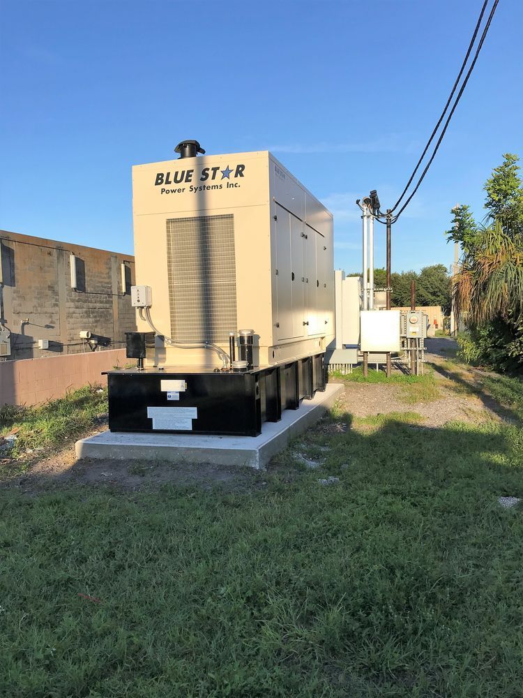 Large beige industrial generator on a black platform in a grassy lot beside utility poles and buildings.