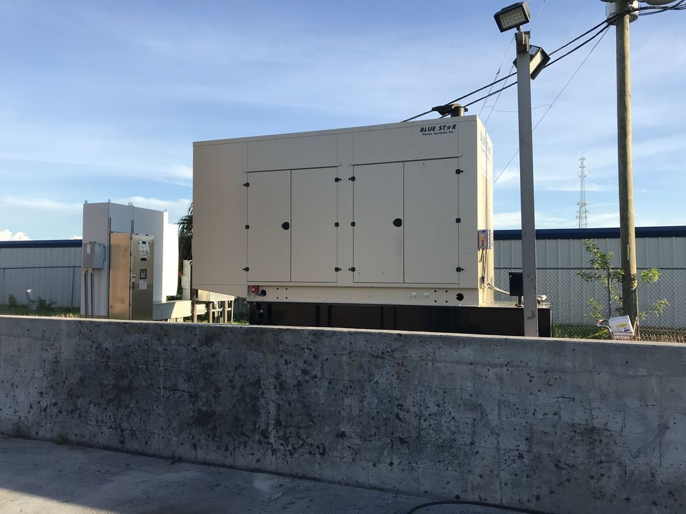 Industrial facility with large white equipment units behind a concrete wall under a blue sky