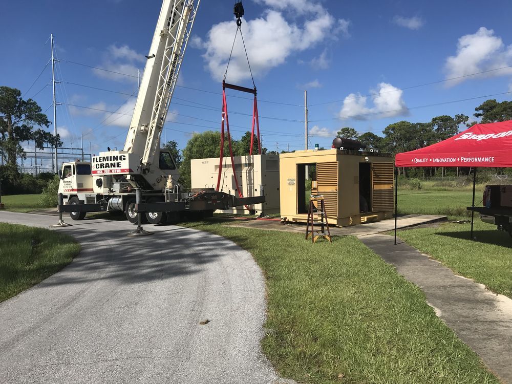 Crane lifting a beige portable building beside a road under a blue sky, with a red tent nearby
