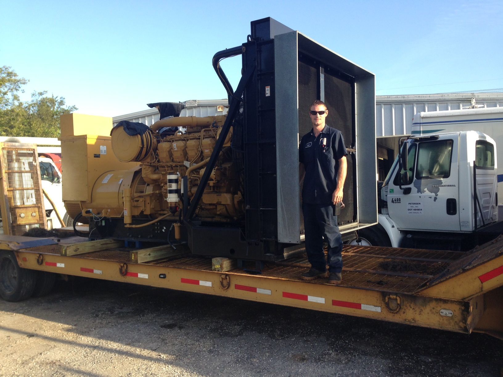 Heavy machinery on a flatbed trailer beside a white truck, with a person standing on the trailer.