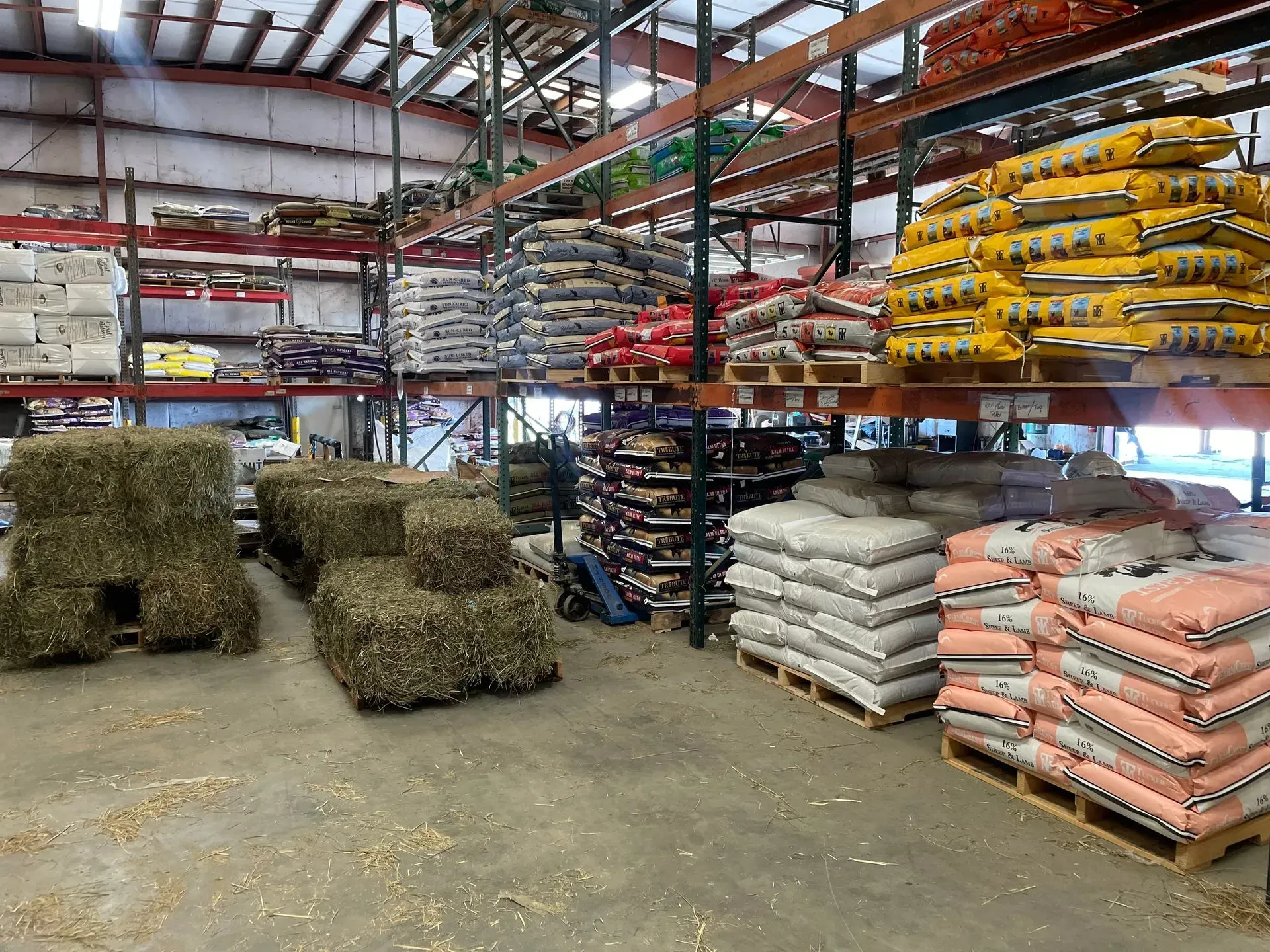 Hay bales and stacked bags of feed in a large warehouse with metal shelves.