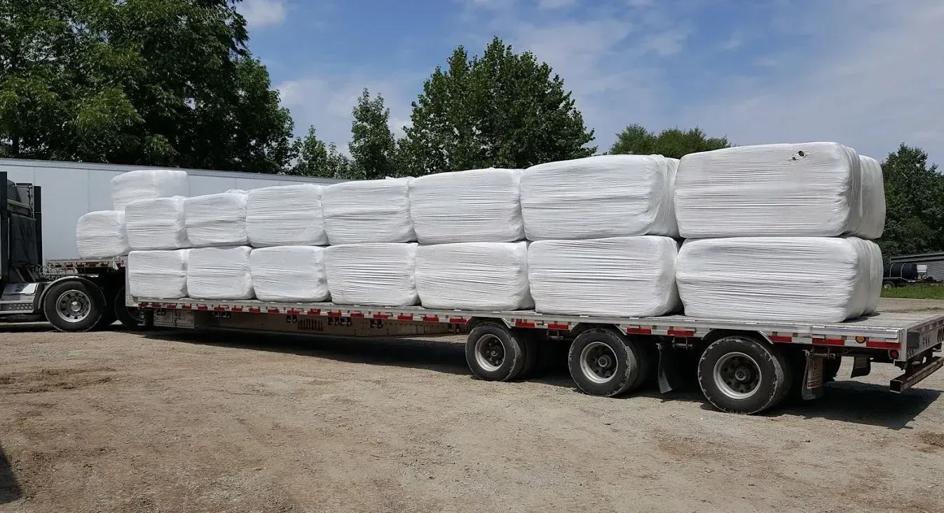 A flatbed truck loaded with multiple large, white, wrapped bales, parked on a gravel lot.