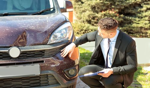 Man crouching beside a maroon car, inspecting the front bumper outdoors in bright sunlight