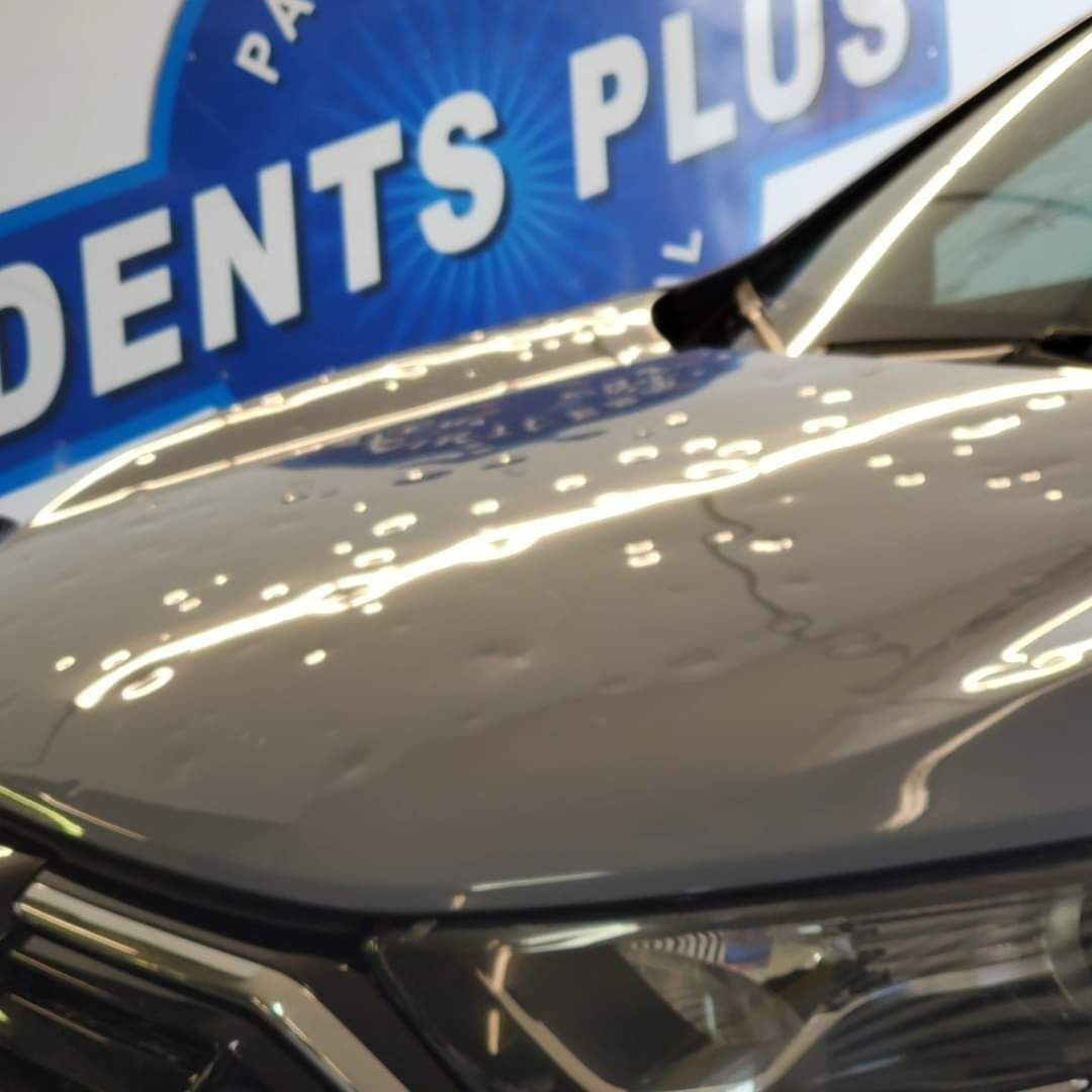 Close-up of a gray car hood and headlight at an auto display with a blue dealership sign in the background