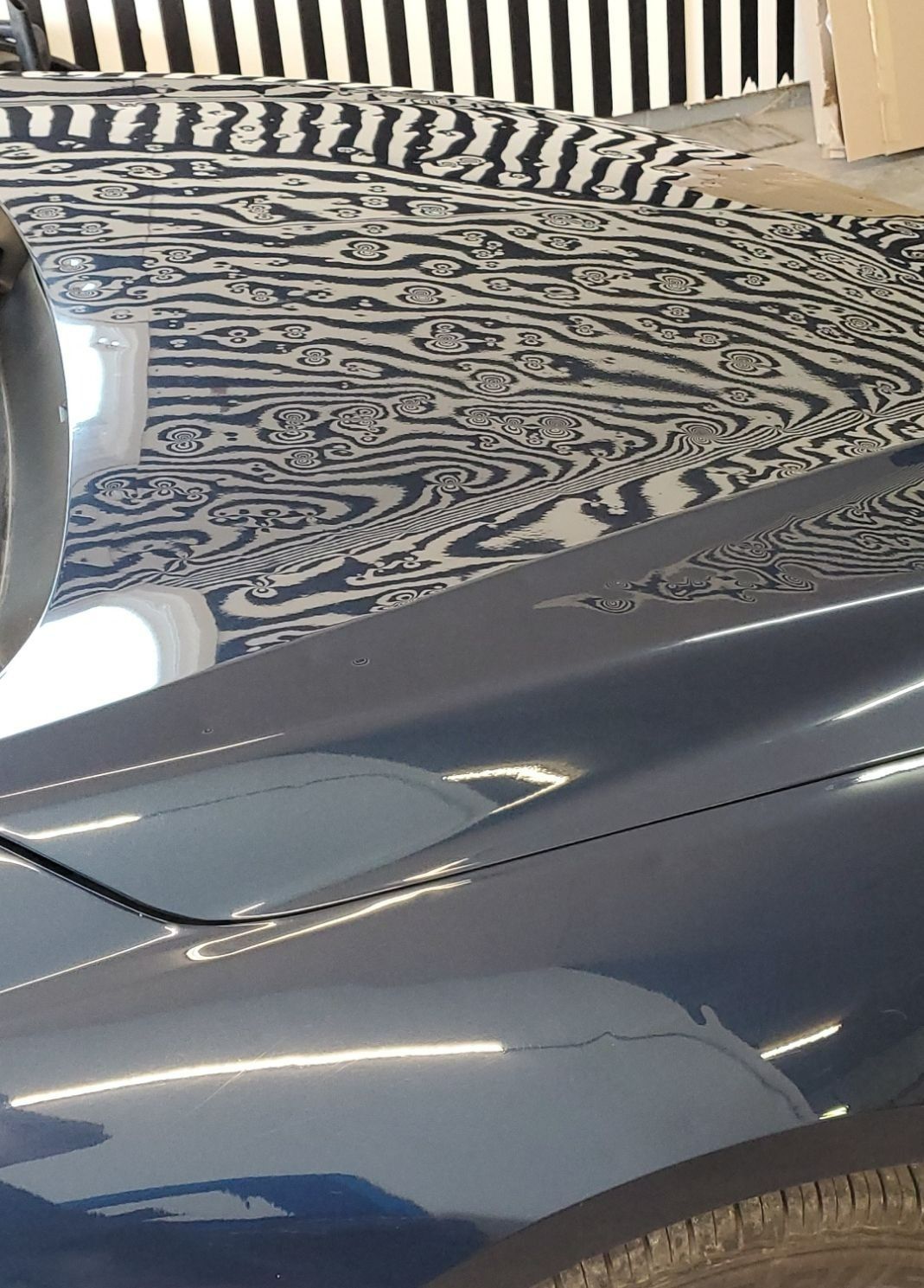 Close-up of a dark blue car hood reflecting a black-and-white striped ceiling and a light source