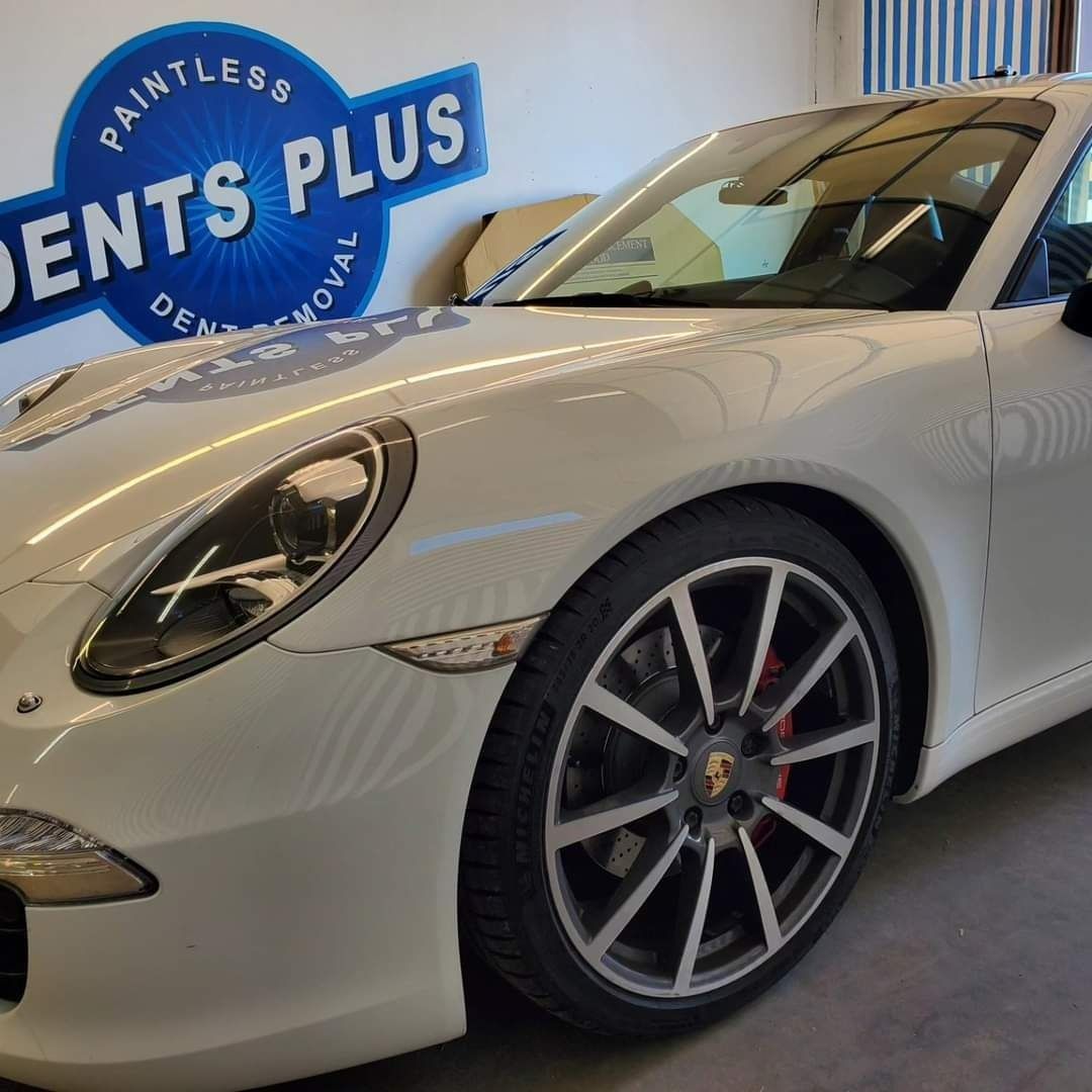 White Porsche 911 parked indoors beside a blue “Dentless Plus” sign.