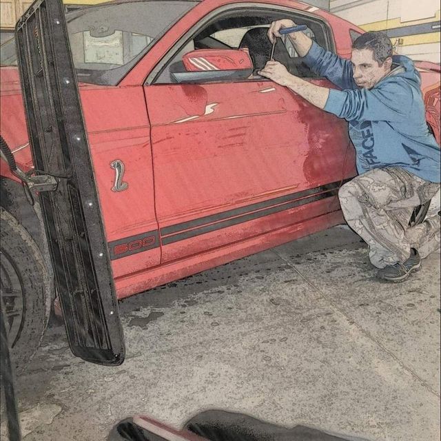 Man crouches beside a red truck, working on the driver-side window in a garage.