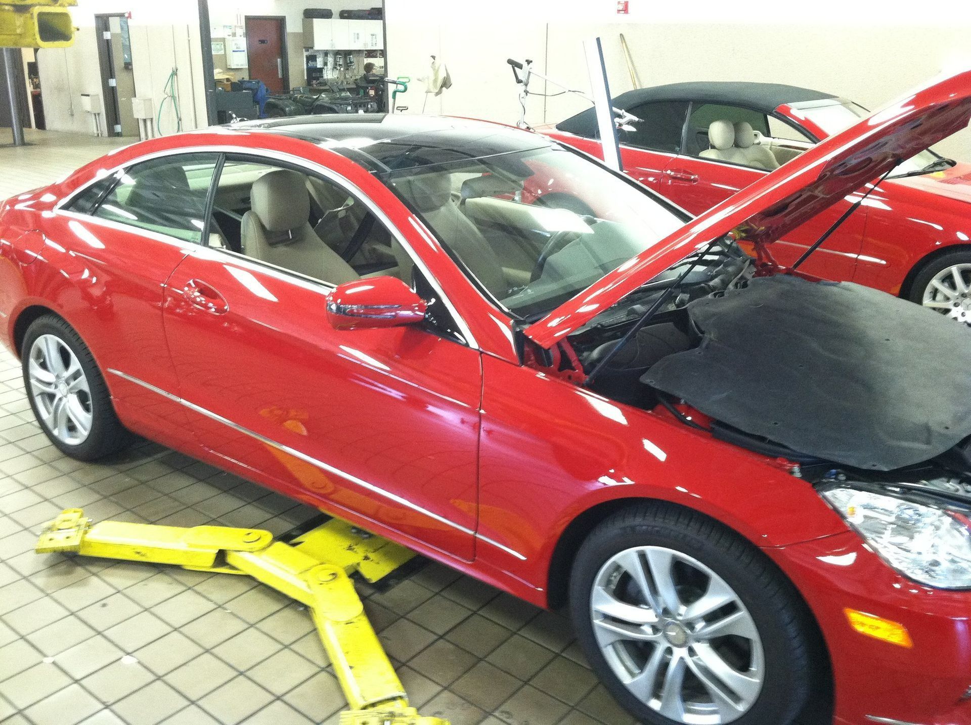 Red car in an auto repair shop with its hood open and a wheel removed on a yellow jack stand.