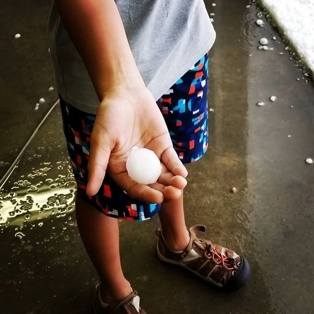Person holding a white ball over a wet pavement, wearing patterned shorts and sandals.