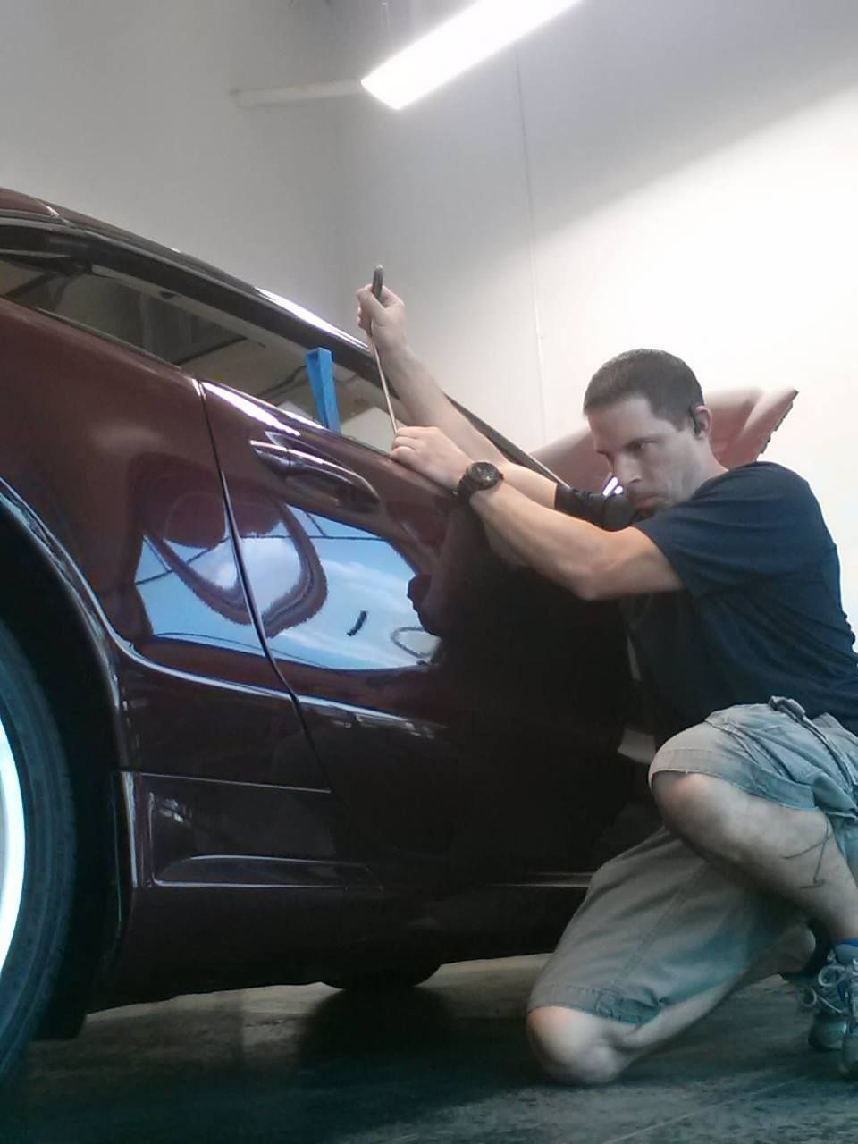 Man kneeling beside a maroon car in a garage, working on the rear panel with a tool.