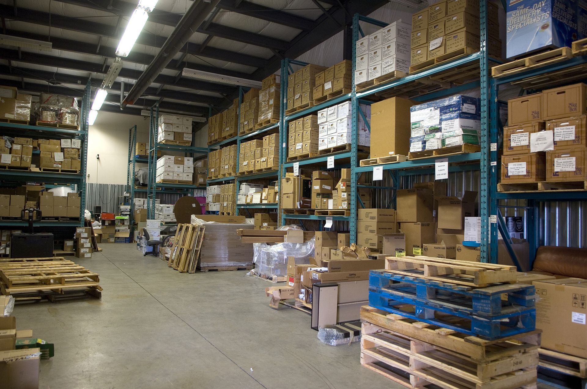 Warehouse aisle with stacked pallets, boxes, and shelves of stored goods under industrial lighting