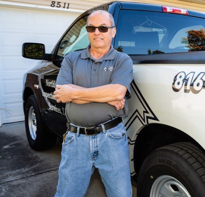 Man standing with arms crossed beside a black-and-white tow truck in a driveway, wearing sunglasses and jeans
